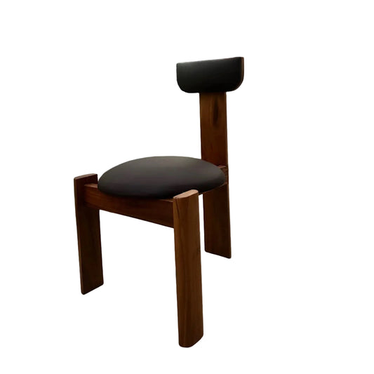 A wooden chair with a dark cushioned seat and backrest, photographed outdoors on a concrete surface with a gate, tree, and building in the background.